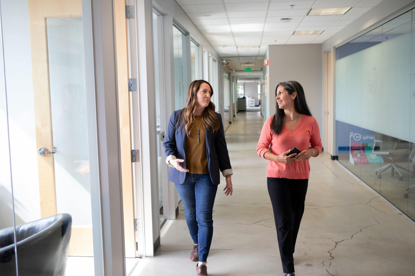 Two professional women engaged in conversation while walking through a modern office corridor.