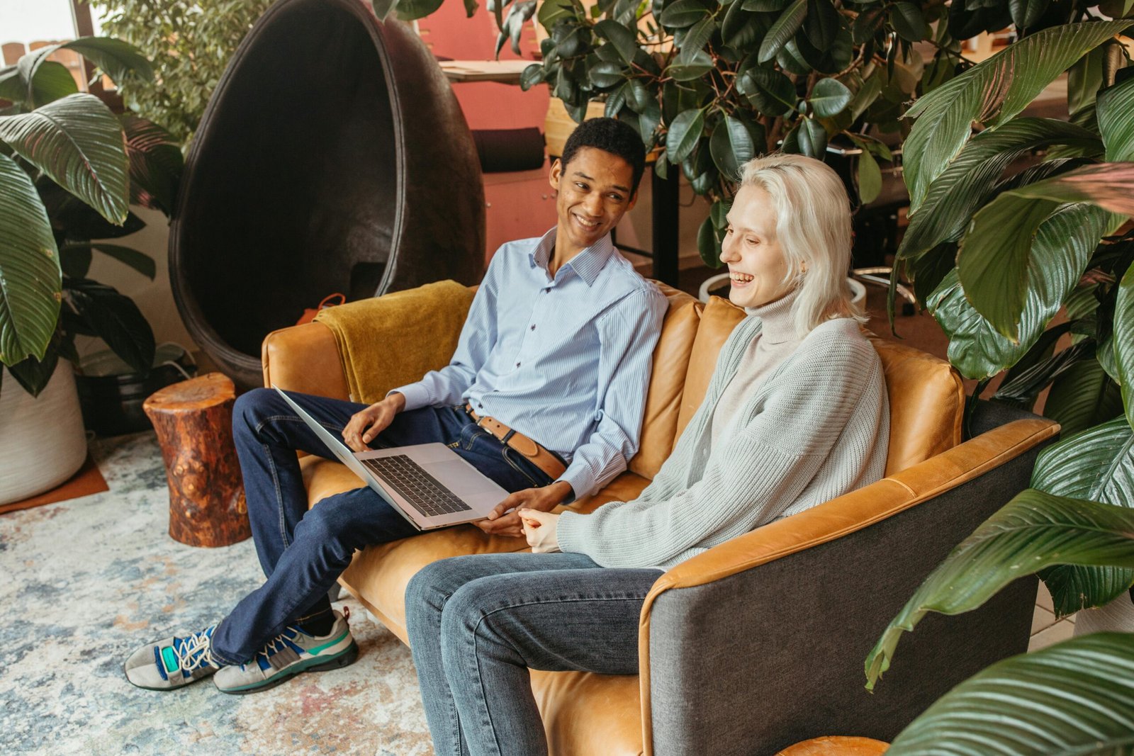 Two colleagues smiling and collaborating on a laptop in a cozy, plant-filled office setting.
