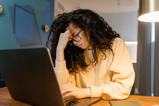 The 5 Most Barely Believable Payroll Disputes of All Time A tired woman with curly hair and glasses experiencing stress while working on a laptop.