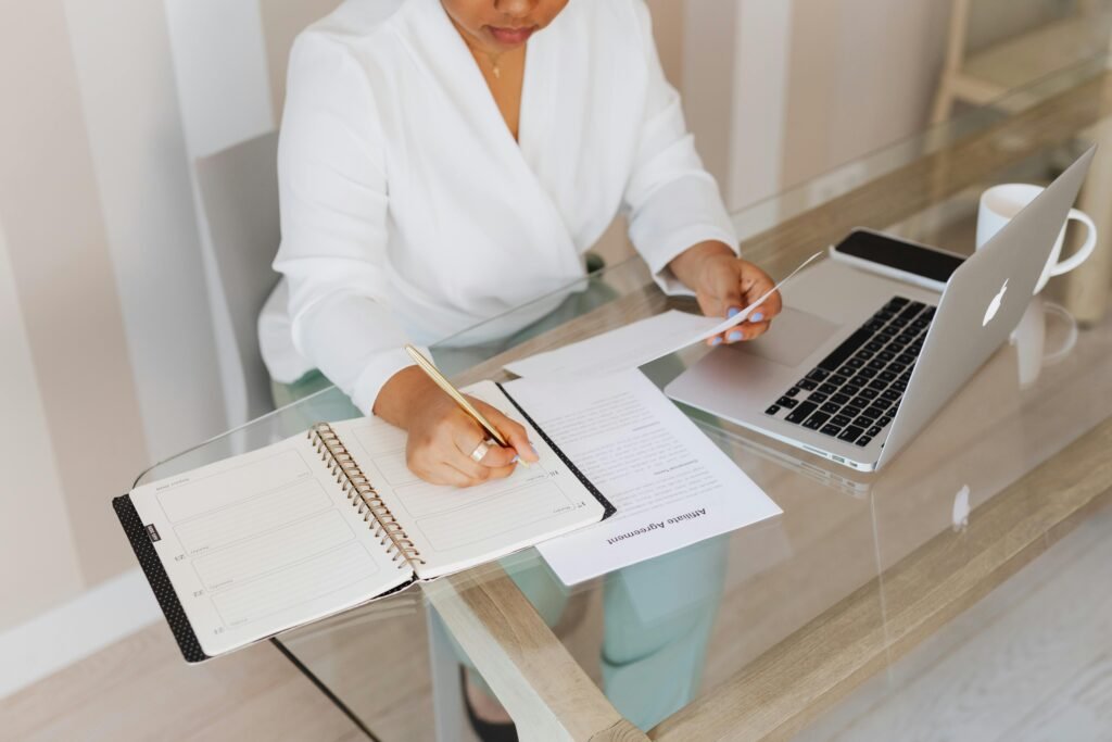 Woman writing notes while working with a laptop and documents at a glass desk indoors.