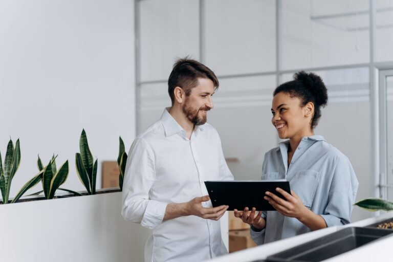 A black woman and a caucasian man discussing work with a clipboard in a modern office setting.